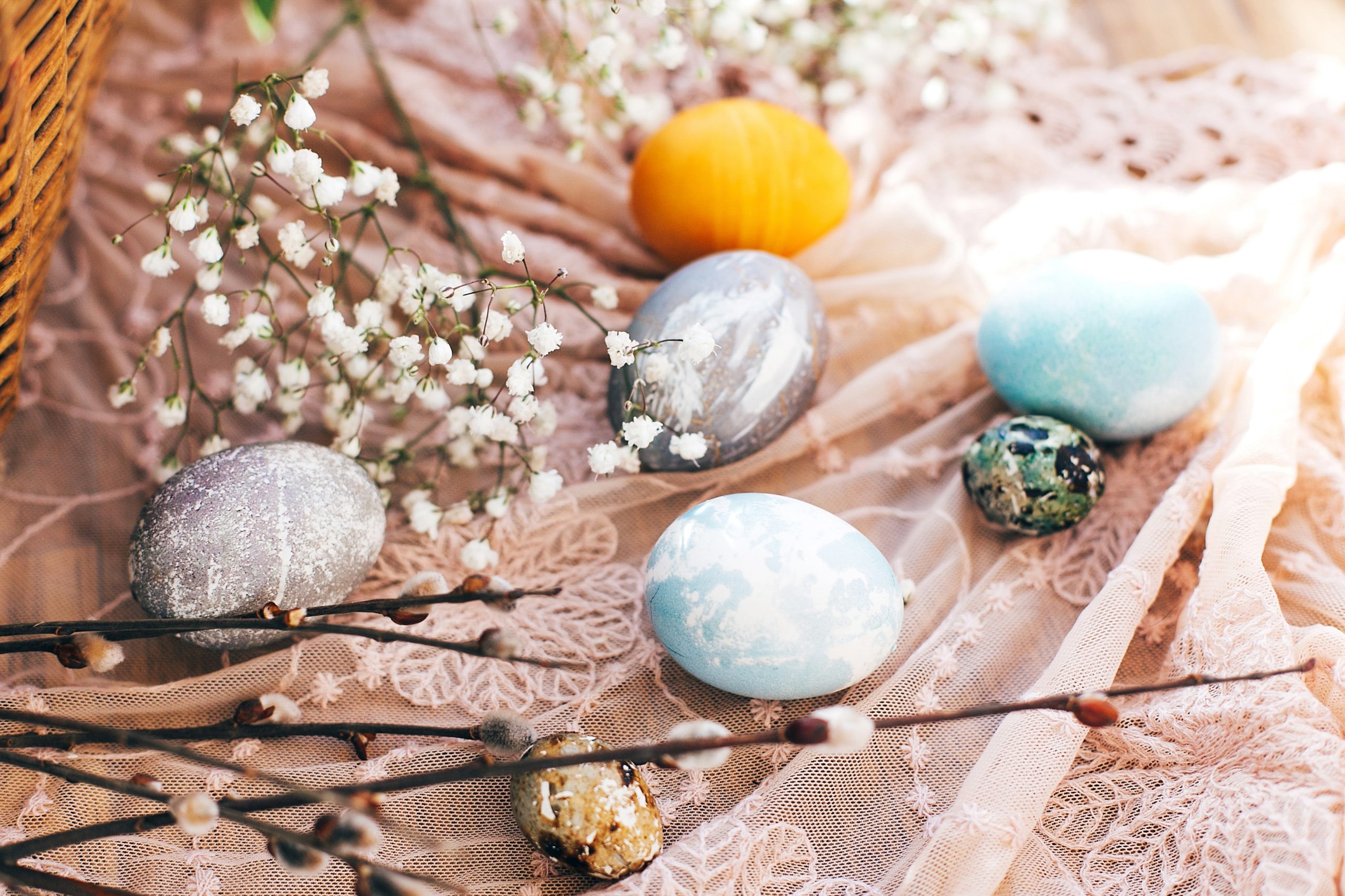 Stylish Easter eggs with spring flowers and willow branches on rustic fabric at wicker basket with holiday food in sunny light. Modern easter eggs painted with natural dye. Happy Easter
